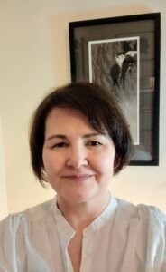 The head and shoulders of a woman with short brown hair and a friendly smile, in front of a farmed black and white picture.